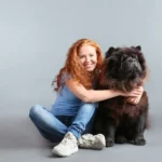 Junge, rothaarige Frau umarmt einen schwarzen Chow-Chow vor einem grauen Hintergrund.